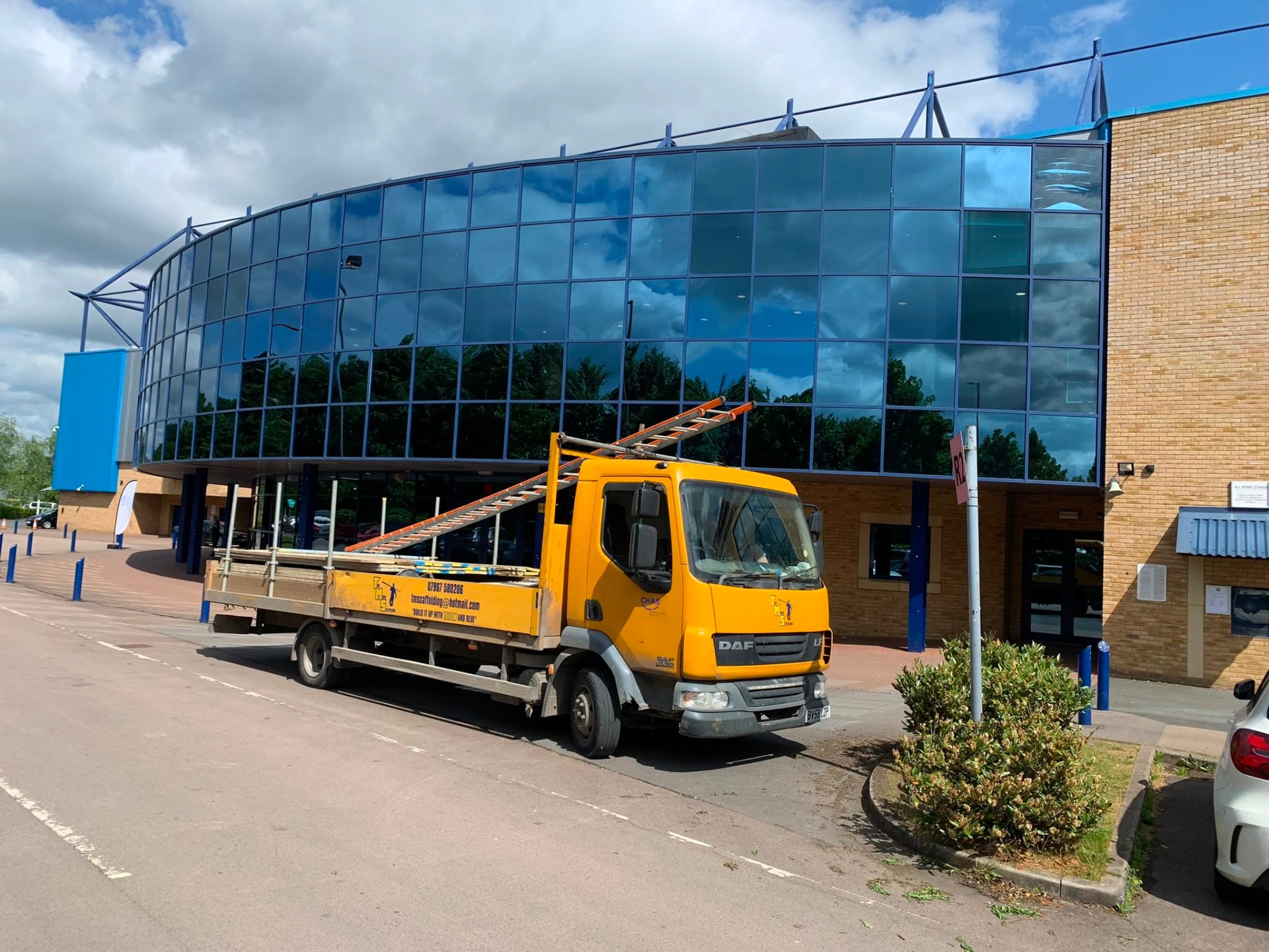 Yellow Tom Moore Scaffolding truck loaded with tube and boards outside a glass-fronted commercial building